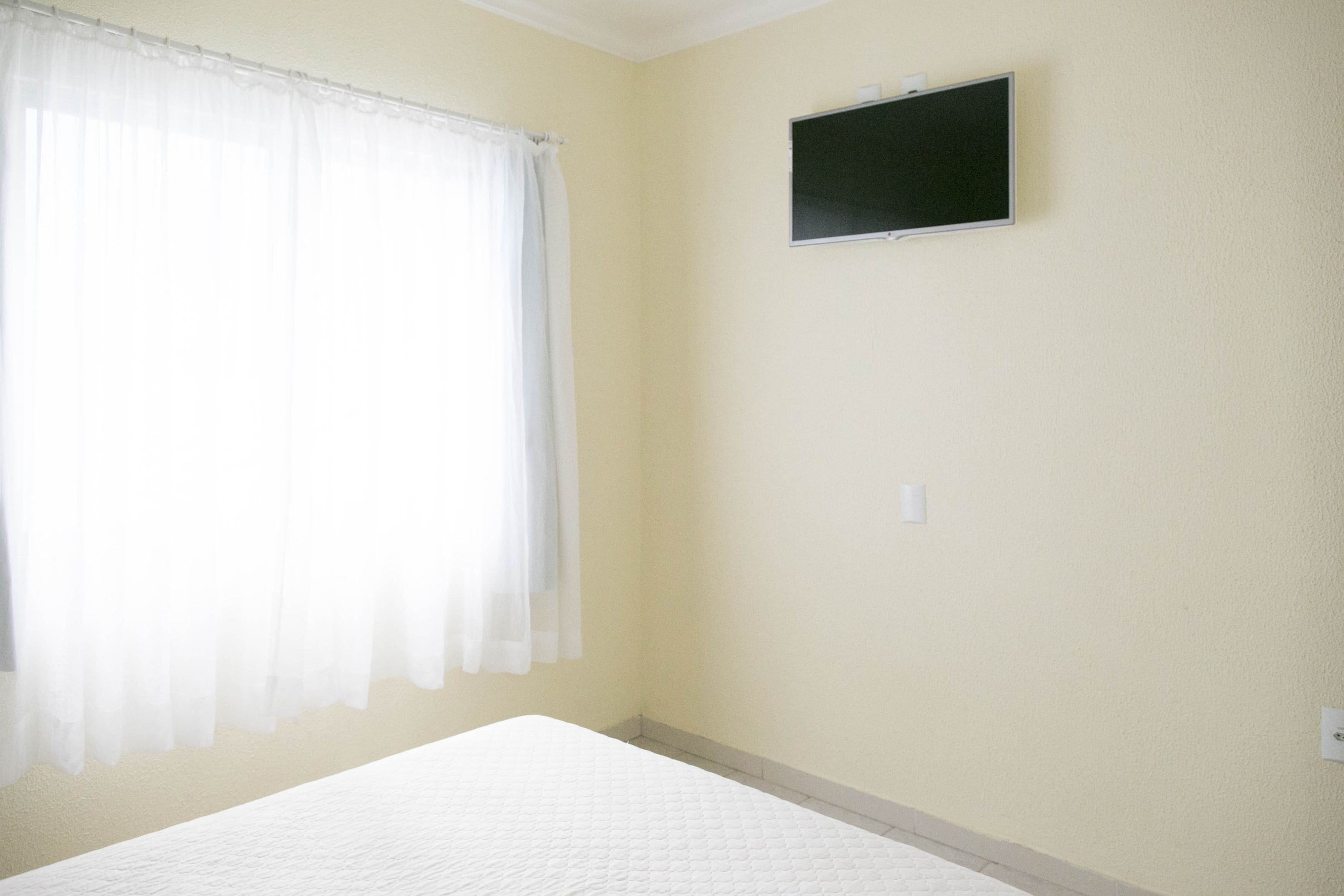 Una habitación con cama, ventana con cortinas traslúcidas, televisor montado en la pared y un interruptor de luz.