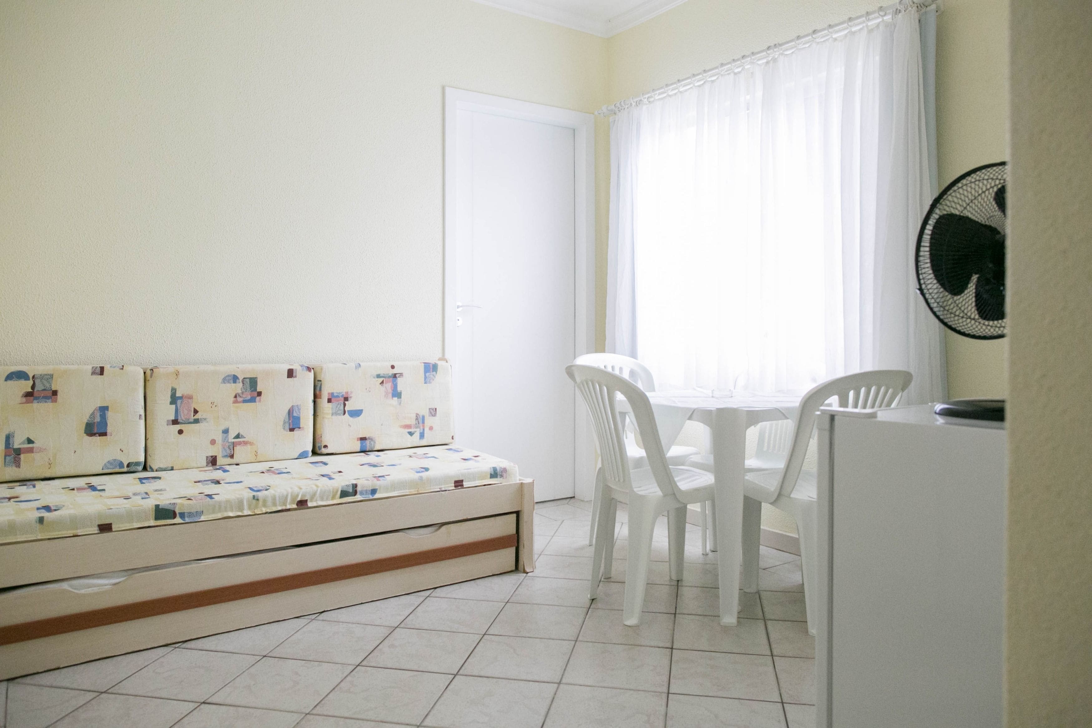 A small, bright room with a white sofa, a dining table with chairs, and a white refrigerator.