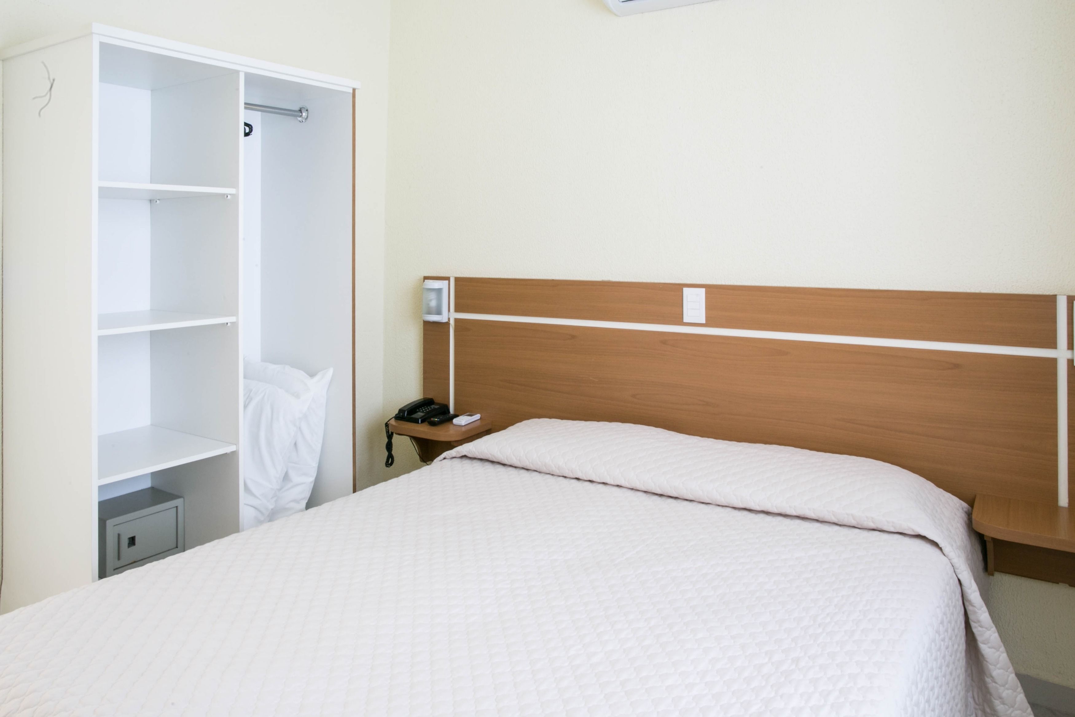 A hotel room with a bed, a wooden headboard, a white wardrobe, and a bedside table with a phone.