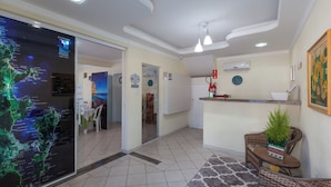 Hall d'albergo con cartellone esplicativo, reception e area relax con una pianta.