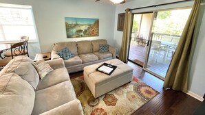A living room with a beige sectional sofa, a white ottoman, a ceiling fan, a painting of a cityscape, and a sliding glass door leading to an outdoor deck. - Beautiful Branson Getaway! (Branson)
