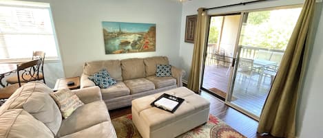 A living room with a beige sectional sofa, a white ottoman, a ceiling fan, a painting of a cityscape, and a sliding glass door leading to an outdoor deck.