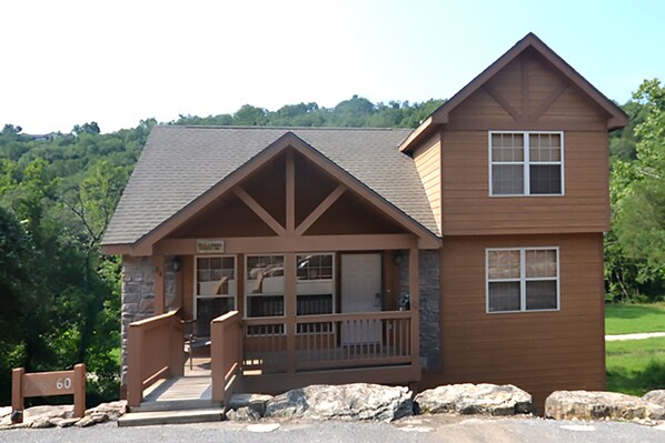 A two-story lodge with a stone facade, a wooden porch, and a clear view of the surrounding forest.
