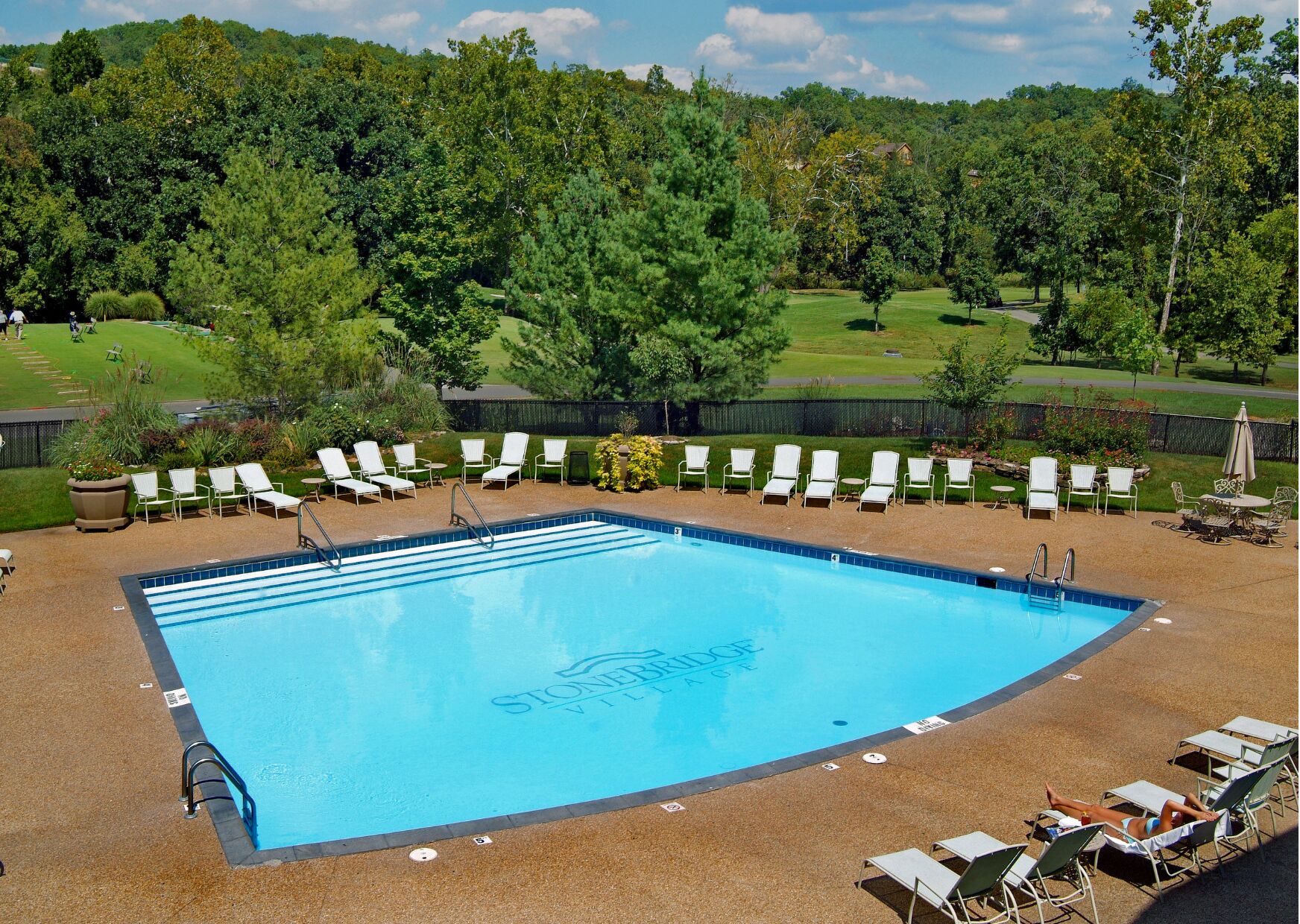 A rectangular swimming pool with a blue interior, surrounded by white lounge chairs and a paved deck.