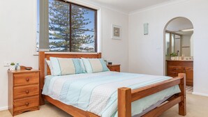 Ein Schlafzimmer mit einem hölzernen Bett, einem Nachttisch, einer Kommode, einem großen Fenster mit Blick auf Bäume und einem Spiegel im Badezimmer.