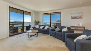 Ein großzügiges Wohnzimmer mit einer großen, grauen Sofagarnitur, einem runden Couchtisch und Meerblick durch große Fenster.
