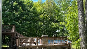 A wooden deck with a staircase leading to a covered area, surrounded by lush greenery and trees. - Heated pool-fire pit-central air-near PRNL (Christmas)