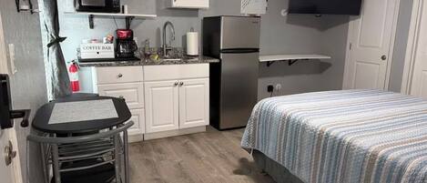 A compact kitchen with a microwave, coffee maker, and refrigerator. There is a small dining table and a bed with a striped bedspread.