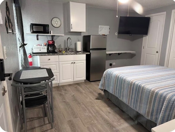 A compact kitchen with a microwave, coffee maker, and refrigerator. There is a small dining table and a bed with a striped bedspread.