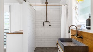 Une salle de bain avec des murs recouverts de carreaux blancs, un meuble-lavabo en bois, une douche dotée d’un pommeau de douche incurvé et un porte-serviettes avec des serviettes.