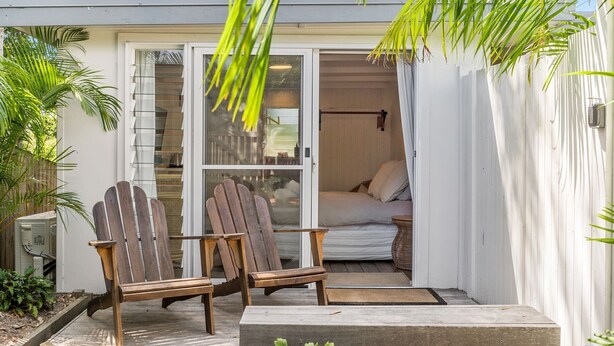 Un espace extérieur accueillant, aménagé avec deux fauteuils Adirondack en bois, une petite table et une porte-fenêtre coulissante donnant sur une chambre à coucher équipée d’un lit et d’oreillers.