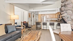 Una sala de estar moderna con un sofá, sillas, una mesa de centro y una zona de cocina con armarios de madera y una pared de piedra.