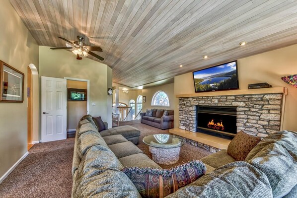 A spacious living room with a stone fireplace, a large flat-screen TV, and a ceiling fan.