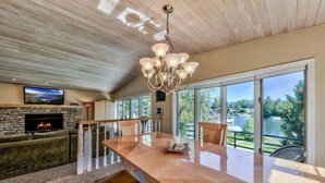 Un salón comedor espacioso con una gran mesa de madera, chimenea y vistas a un lago a través de amplios ventanales.