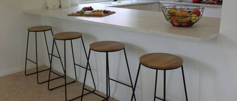 A kitchen with a white countertop, three wooden stools, a fruit bowl, and a potted plant.