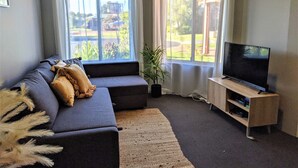 Ett vardagsrum med en soffa i mörk färg, en platt-TV på ett träställ och stora fönster med vita gardiner.