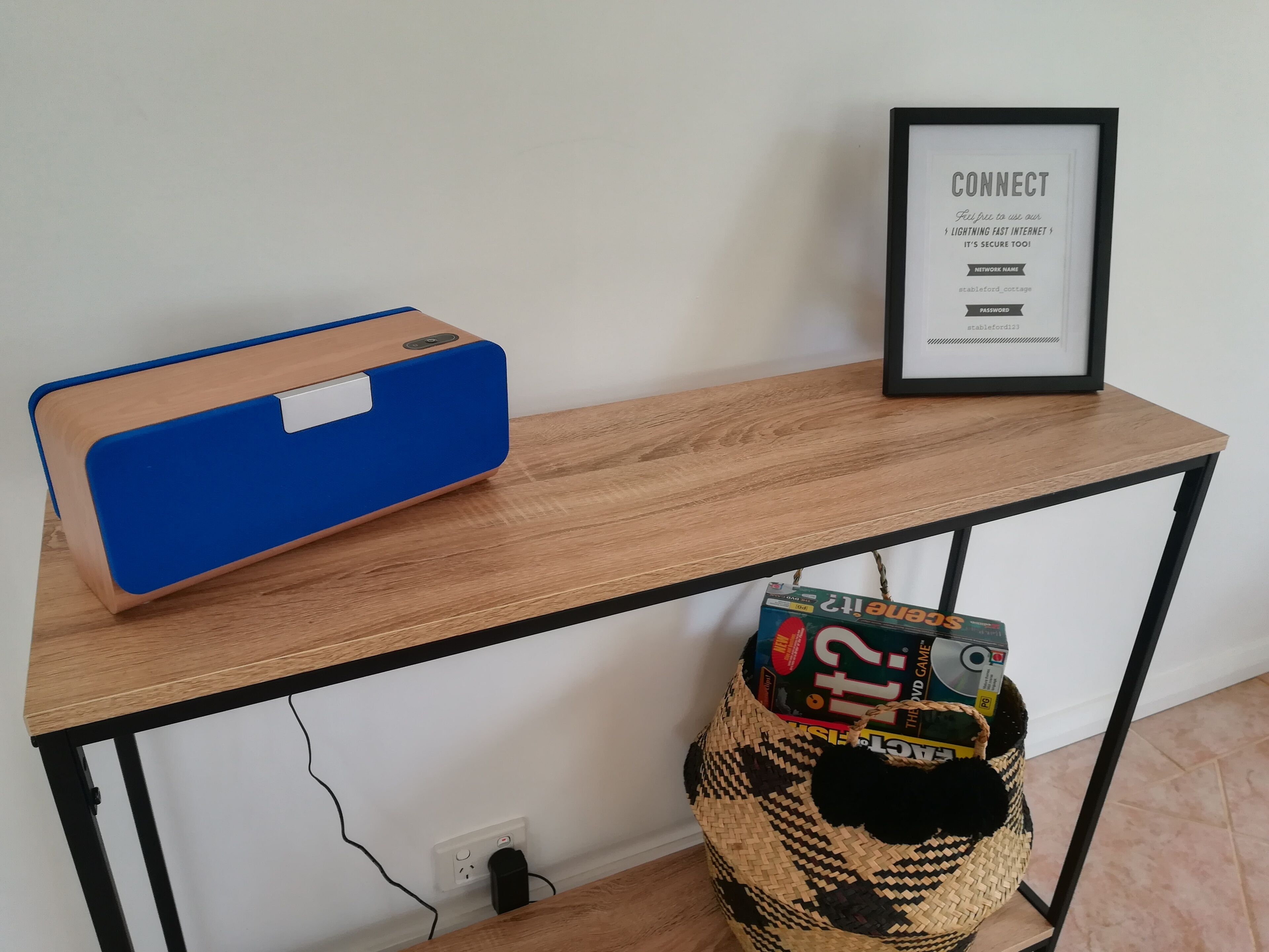 A wooden table with a blue speaker, a framed sign, and a woven basket with a DVD game box.