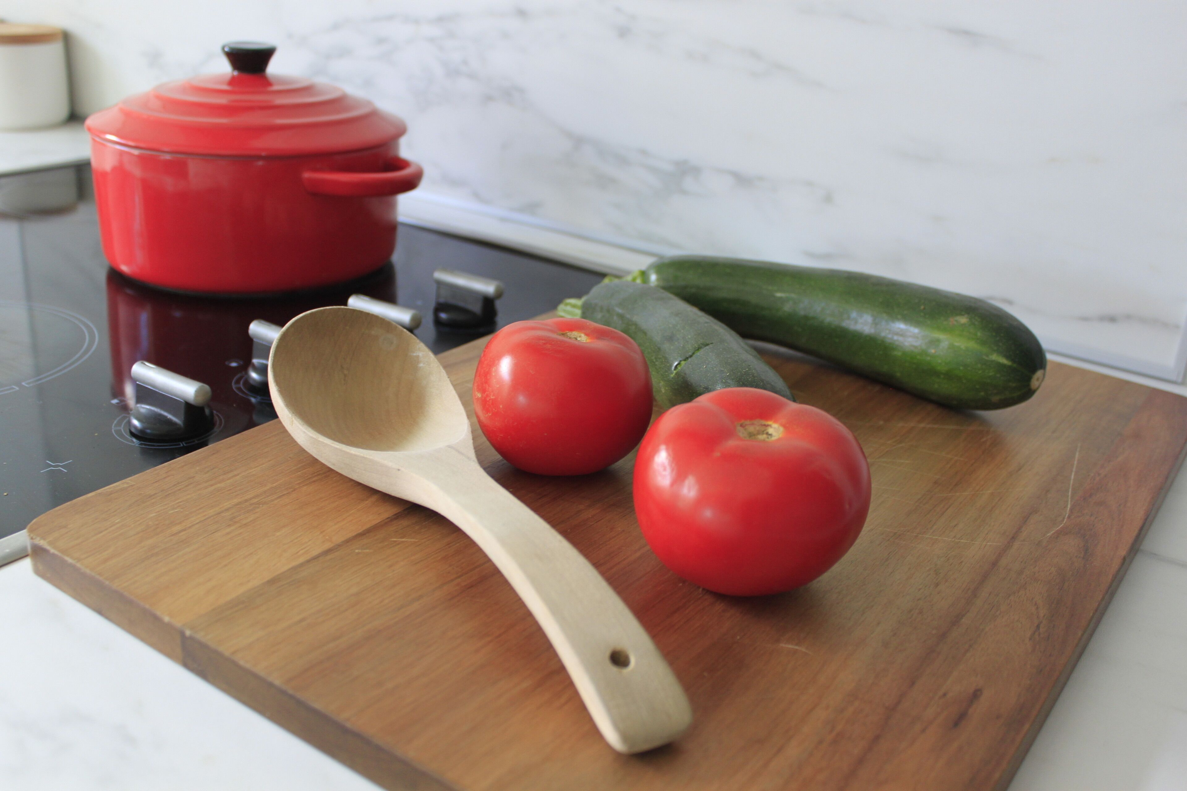 A kitchen counter with a red pot, a wooden spoon, two tomatoes, and a zucchini.