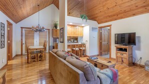 A spacious living area with a wooden ceiling, a dining table with chairs, a sofa, a television, and a kitchen area with wooden cabinets and a ceiling fan.