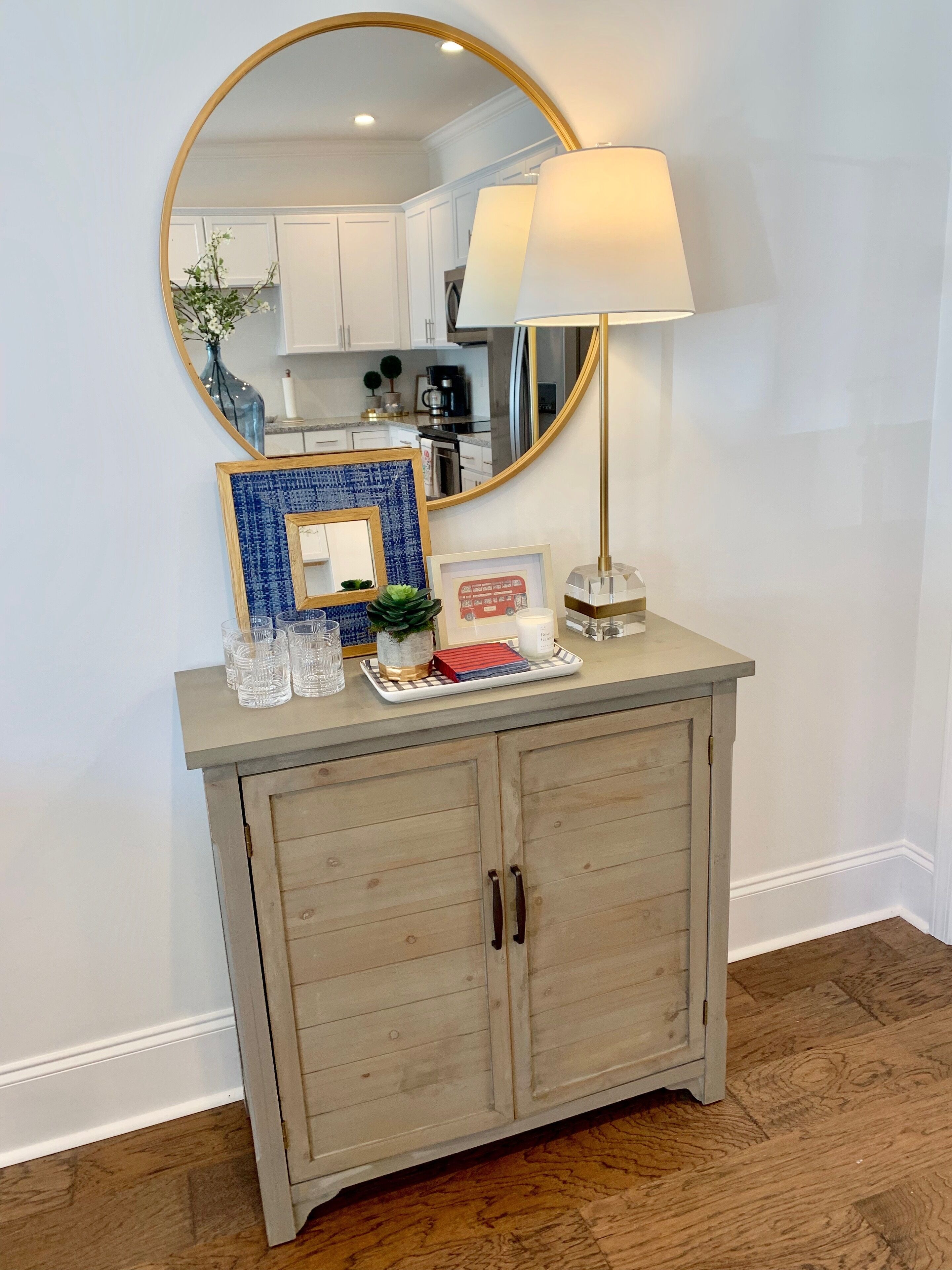 Een keuken met witte kastjes, een ronde spiegel, een lamp en een kleine tafel met glazen en een fotolijst.