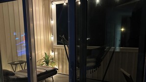 A room with a sliding glass door leading to an outdoor deck. Inside, there is a bed with a striped blanket, a small table with a lamp, and a chair. The deck has a wooden floor and is equipped with outdoor lighting. - 1-Bedroom Condo Just 1 Block from the Beach and Steps from the Hampton Strip (Hampton)