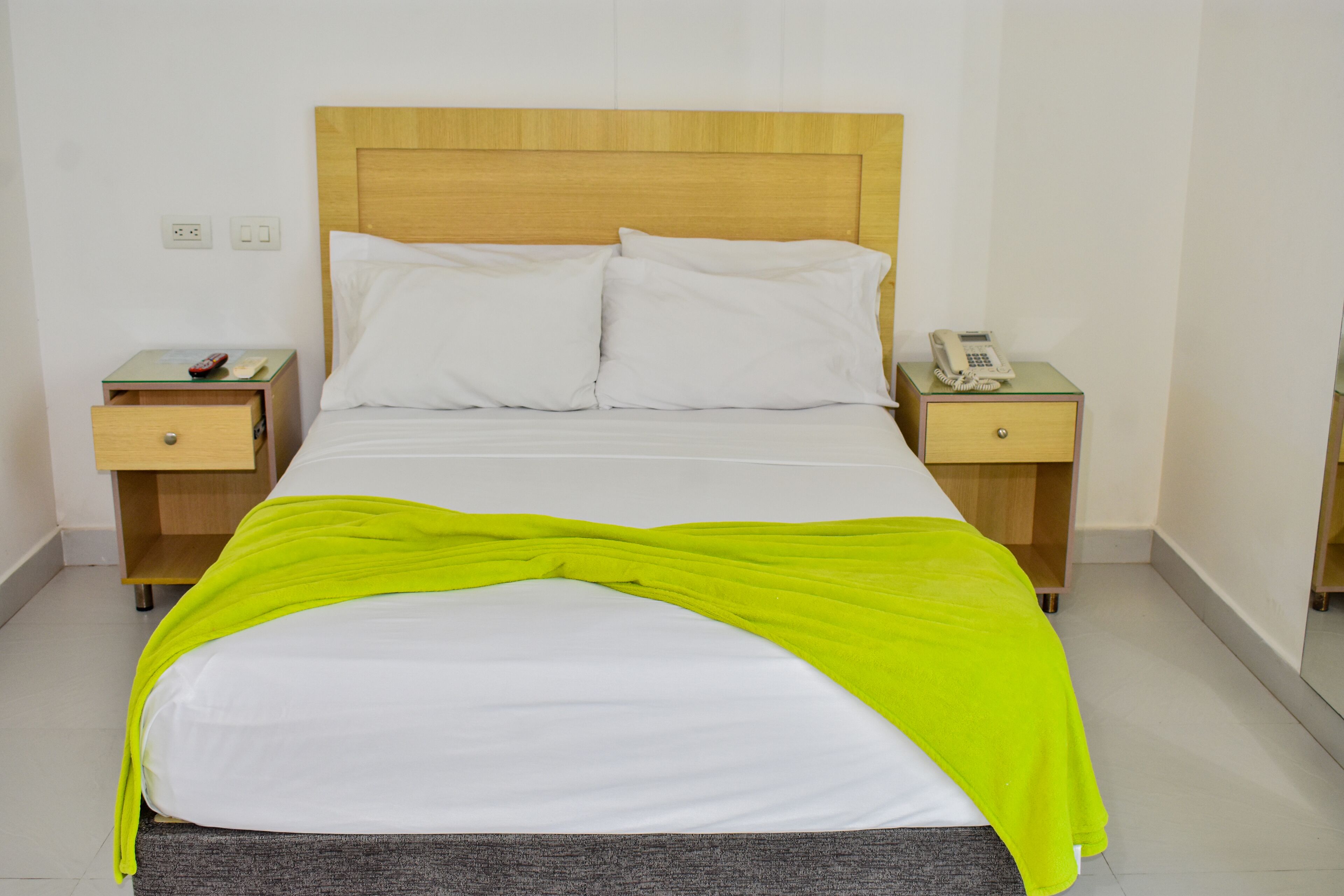 Una cama bien hecha con cabecera de madera, dos mesitas de noche, una con teléfono, y una manta verde brillante.