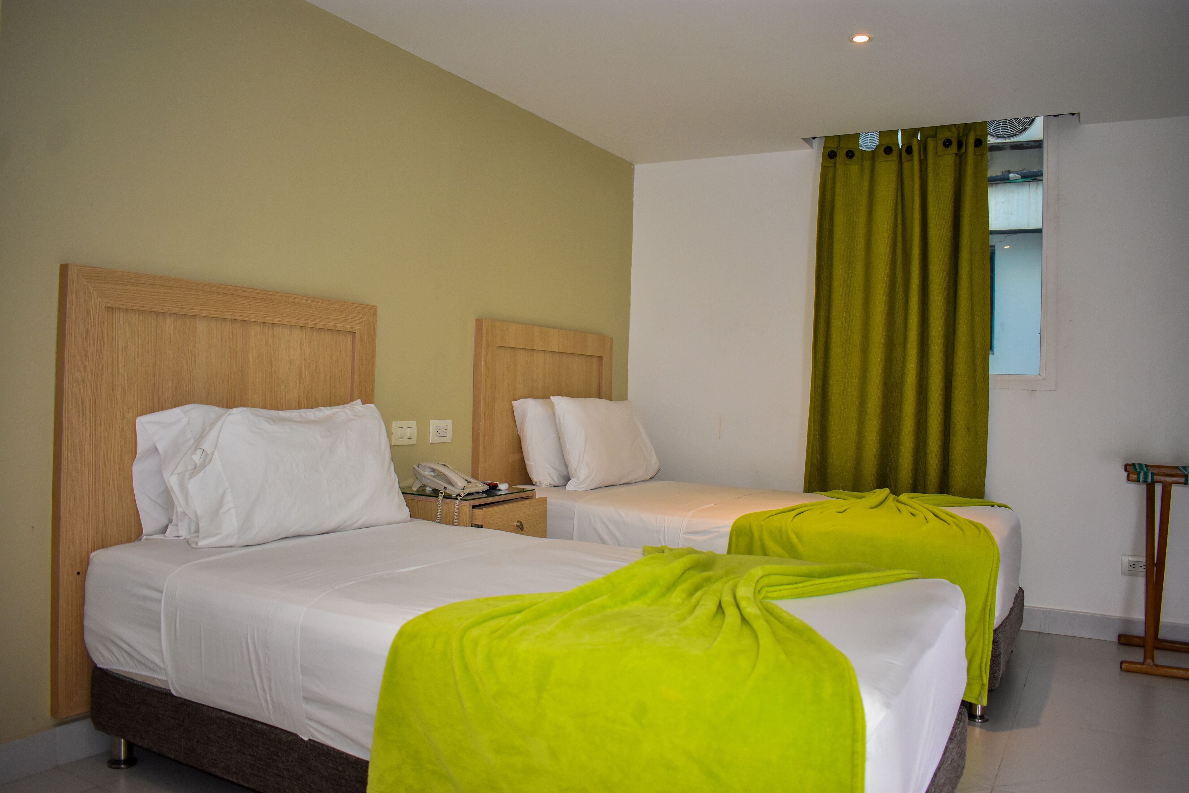 A hotel room with two beds, a bedside table with a phone, and a window with a green curtain.