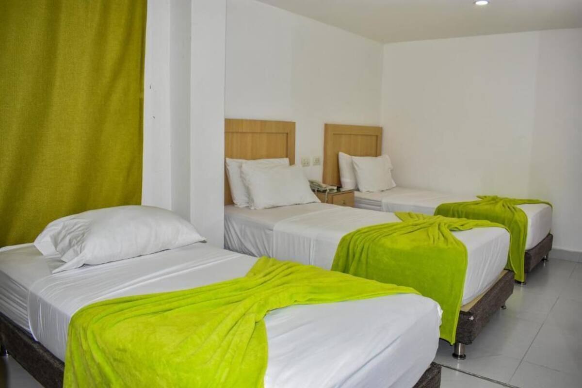 A hotel room with three single beds, each with a white mattress and a green blanket. The room has a wooden headboard and a white wall.