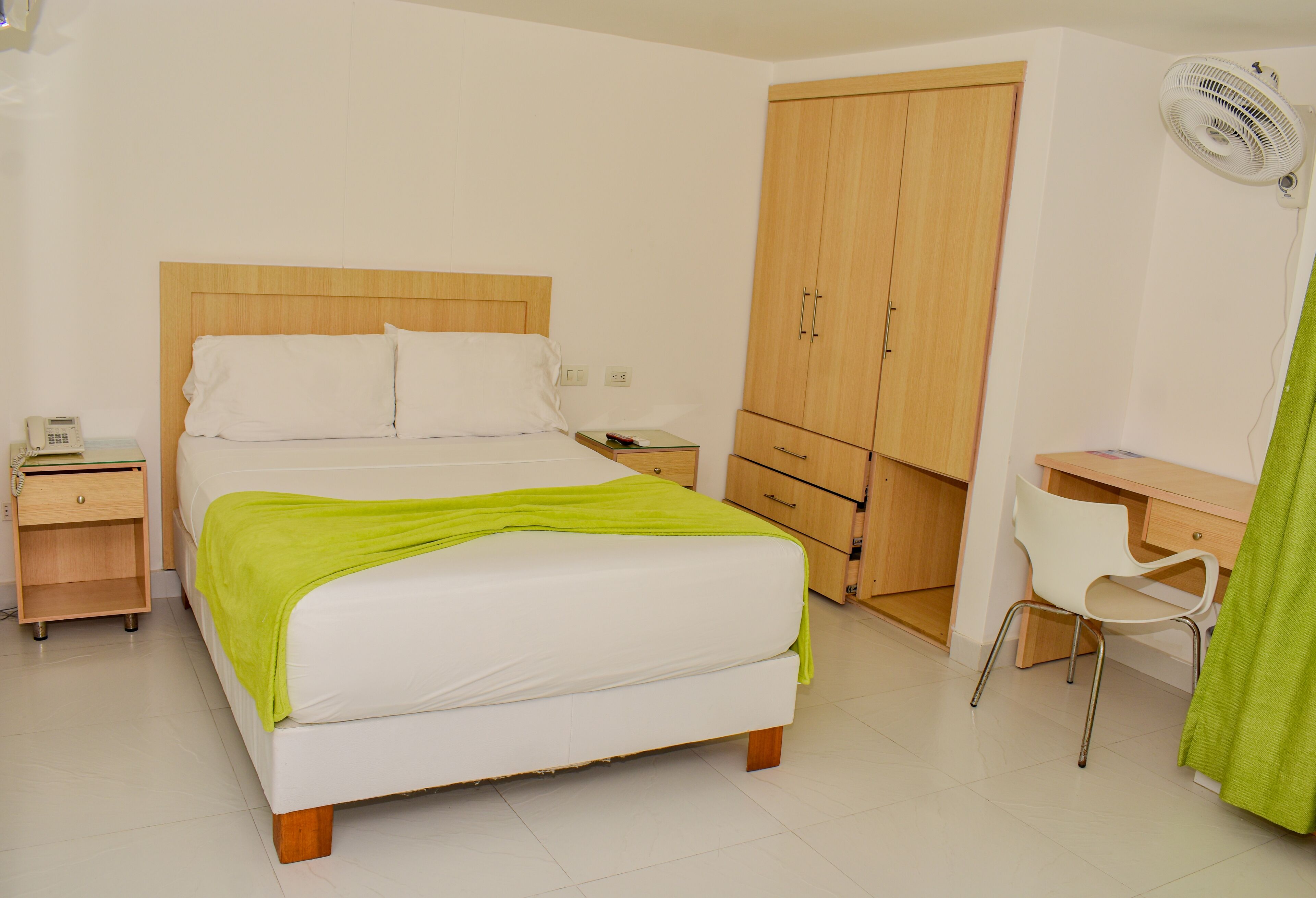 Une chambre à coucher comprenant un lit, des tables de chevet, un bureau, une chaise, une armoire et un ventilateur.