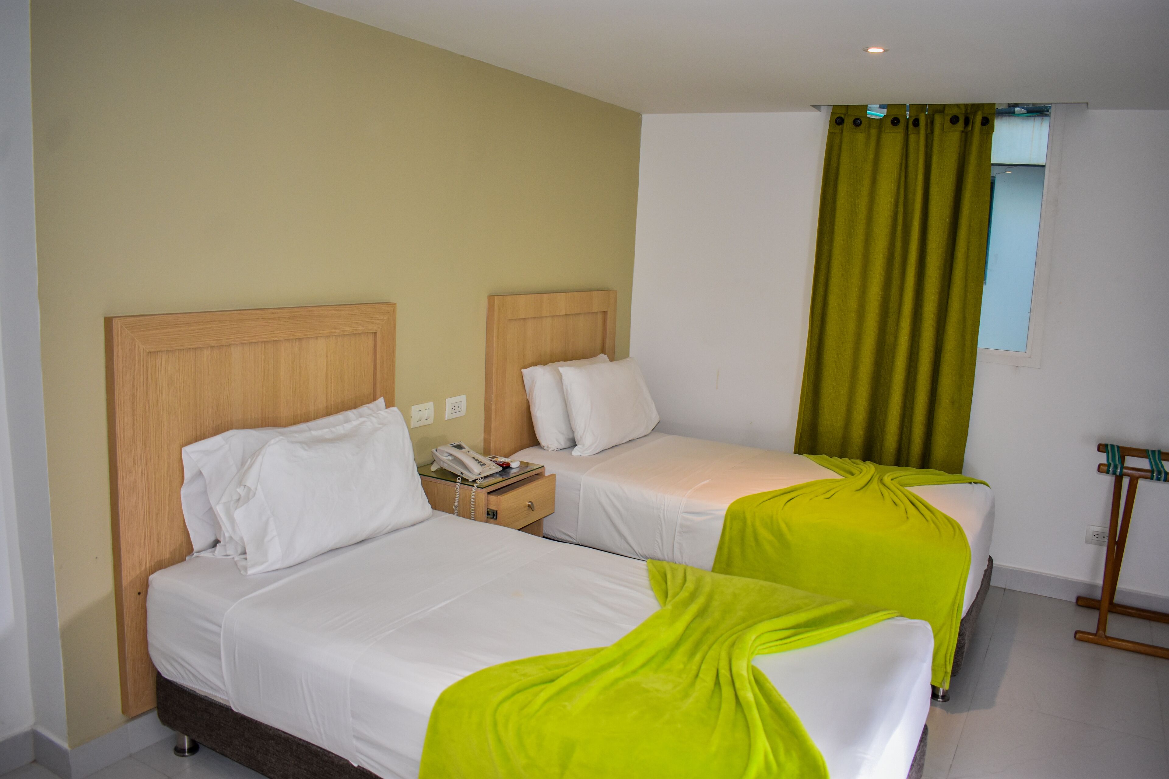 Habitación de hotel con dos camas, cabecero de madera, mesita de noche con teléfono y ventana con cortinas verdes.