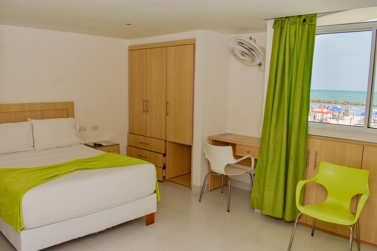 A hotel room with a bed, a desk, a chair, a wardrobe, and a window with a view of the beach.