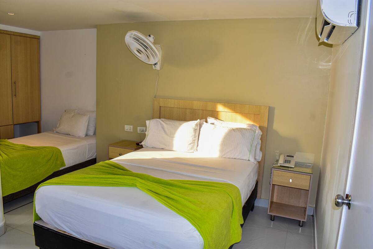 A hotel room with two beds, a bedside table with a phone, and a wall-mounted fan.
