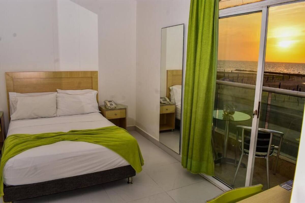 A hotel room with a bed, a bedside table, a mirror, and a balcony with a view of the sunset.