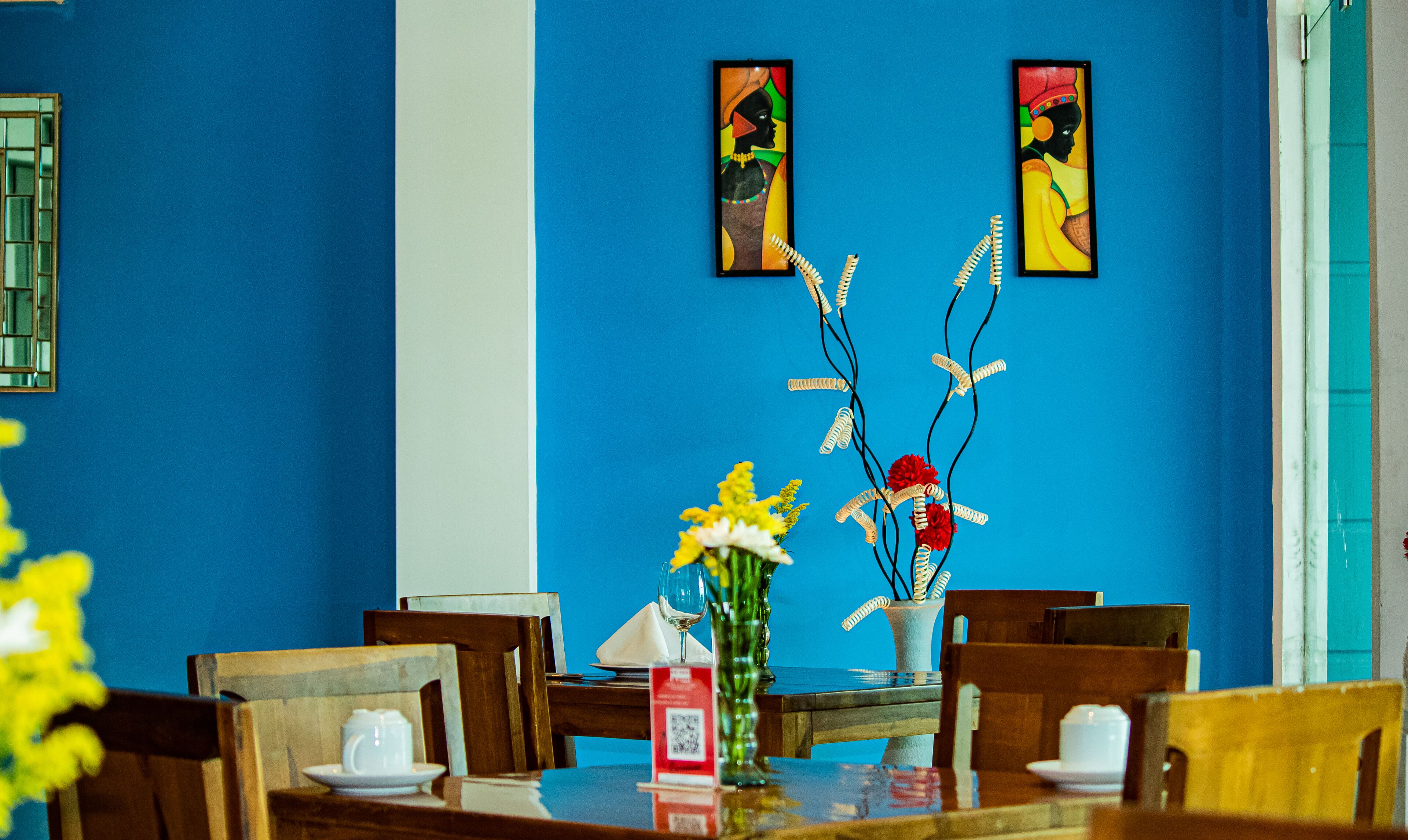 A dining area with a blue wall, two framed artworks, a vase with flowers, and wooden chairs.
