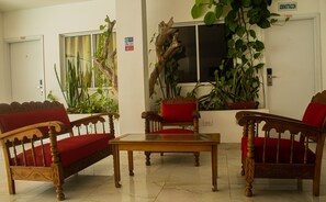A room with a wooden sofa, a wooden table, and a chair with red cushions. There are plants on the windowsill and a sign on the wall.