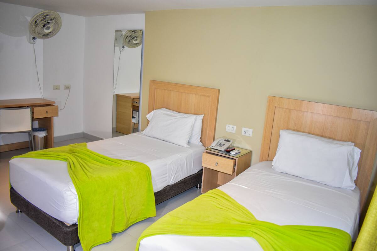 A hotel room with two beds, each with a wooden headboard, white bedding, and a bright green throw blanket. There is a bedside table with a telephone and a wall-mounted fan.