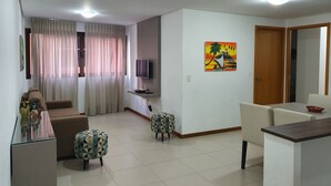 A modern living room with a flat-screen TV mounted on the wall, a dining area with a table and chairs, and a painting of a tropical scene. - Rosa Branca apartments (Ponta Verde)