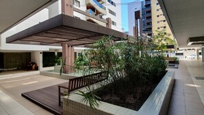 A modern urban plaza with a tiled floor, a wooden bench, a planter with greenery, and a view of high-rise buildings under a clear sky. - Rosa Branca apartments (Ponta Verde)