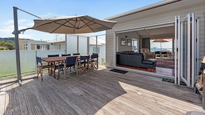 A wooden deck with a table and chairs, an umbrella, and a sliding glass door leading to a living area with a sofa and a view of the ocean. - Sea Shells By The Shaw  - Waihi Beach Holiday Home (Waihi Beach)
