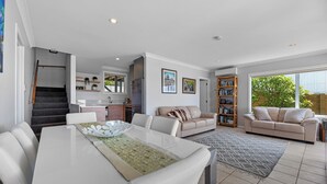 A modern living room with a dining area, a staircase, a kitchen, and a large window with a view of greenery. - Sea Shells By The Shaw  - Waihi Beach Holiday Home (Waihi Beach)