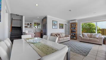 A modern living room with a dining area, a staircase, a kitchen, and a large window with a view of greenery.