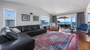 A modern living room with a black leather sofa, a patterned rug, and a view of the sea through large windows. - Sea Shells By The Shaw  - Waihi Beach Holiday Home (Waihi Beach)