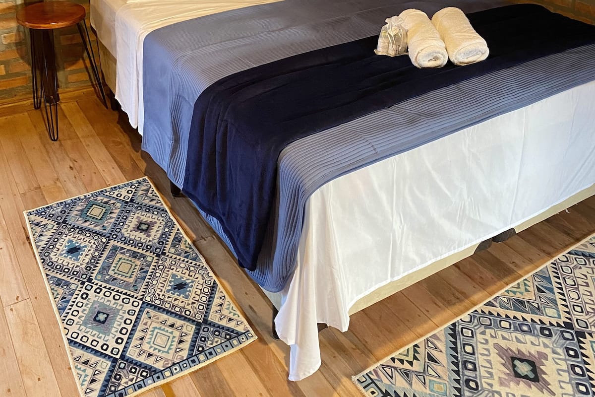 A neatly made bed with a blue and white bedspread, two white pillows, and two rolled towels on the bed. There is a small wooden stool beside the bed and a patterned rug on the wooden floor.