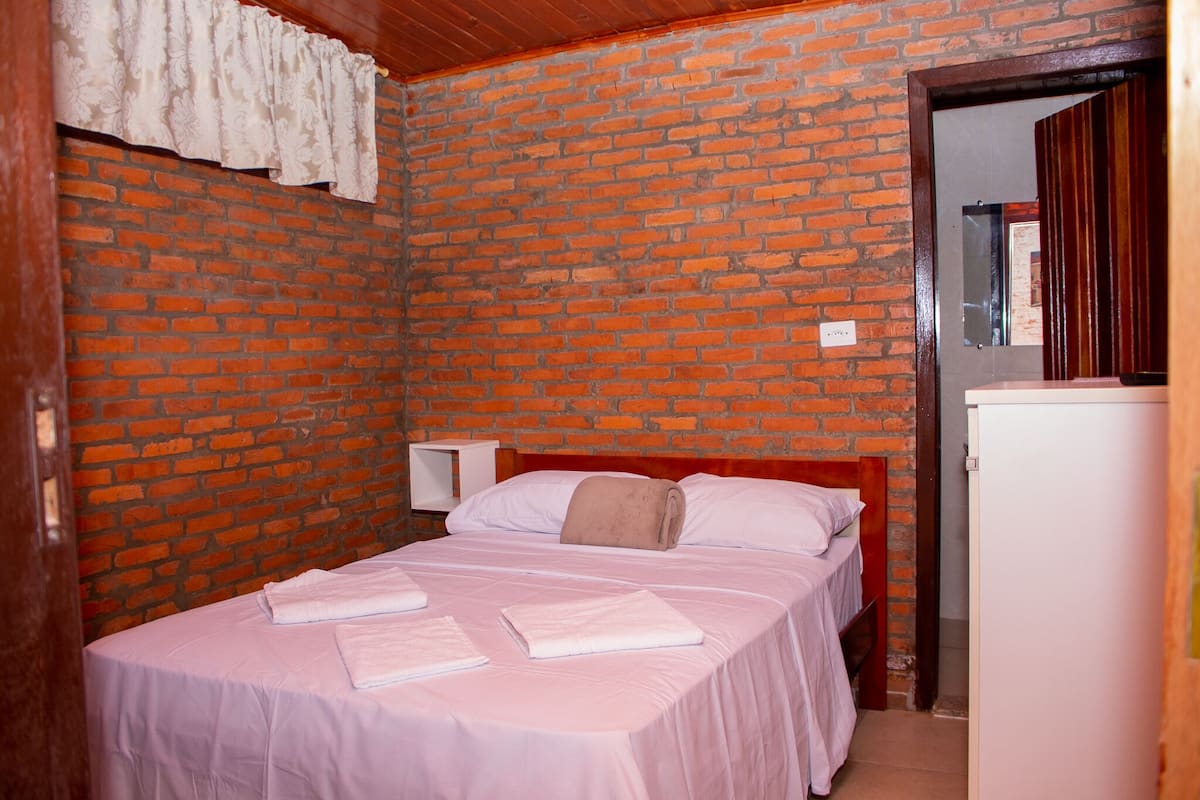 A room with a brick wall, a bed with white linens, a small white nightstand, and a white refrigerator.