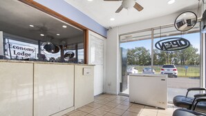 A reception area with a sign reading "Economy Lodge," a ceiling fan, a large window with a view of parked cars, and a reflection of the room in a mirror. - Economy Lodge (Portsmouth)