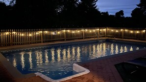 Une piscine entourée d’une clôture en bois et illuminée de guirlandes lumineuses.