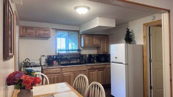 A kitchen with wooden cabinets, a white refrigerator, a dining table with chairs, and a window with blinds.