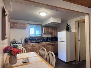 A kitchen with wooden cabinets, a white refrigerator, a dining table with chairs, and a window with blinds.