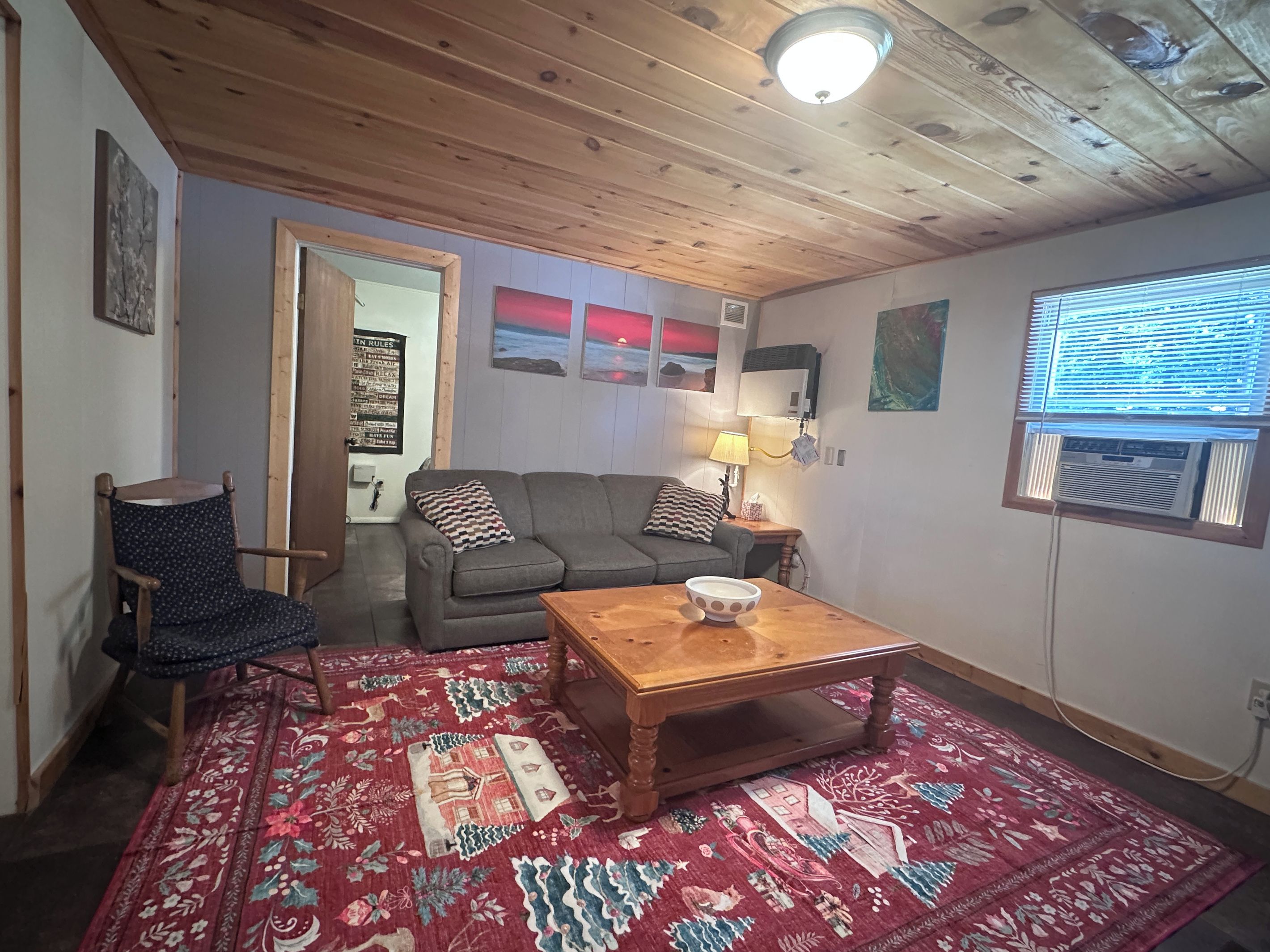 A cozy living room with a wooden ceiling, a grey sofa, a patterned rug, and a wooden coffee table.