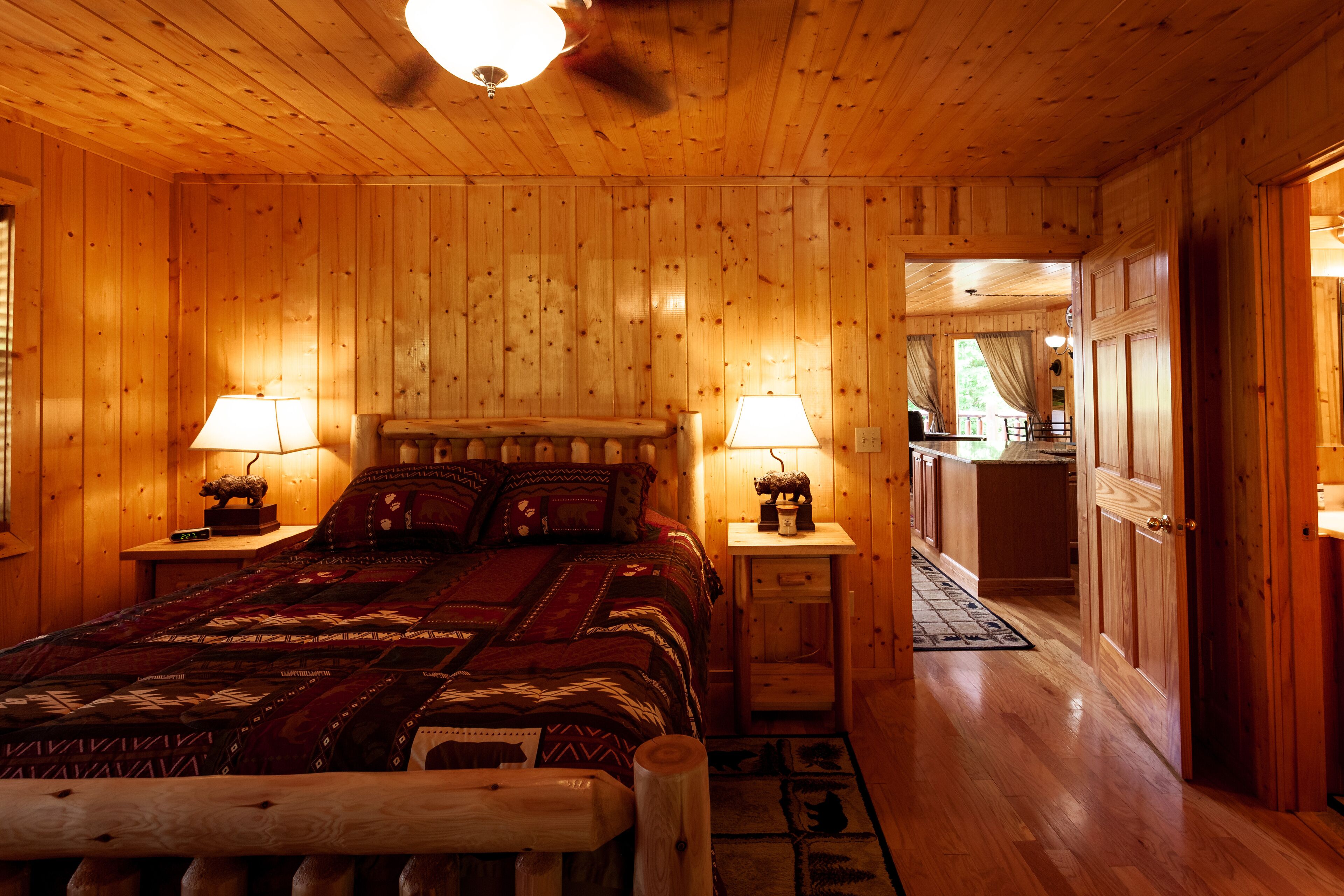 Une chambre aménagée dans une cabane en bois, avec un lit, deux tables de chevet dotées de lampes, une petite table, un banc en bois et une porte donnant sur une autre pièce.